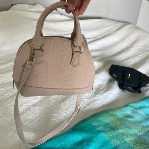 Beige axelväska i mocka-look - Snygg beige axelväska med rundad form och guldfärgade metalldetaljer. Väskan har både handtag och justerbar axelrem, samt dragkedja upptill. Ytan har ett diskret mönster som ger en lyxig känsla. Perfekt storlek för det viktigaste. Man kan ta av axelbandet!