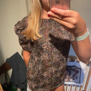 Blommig blus med puffärm - Säljer en blommig blus i mörka toner med korta puffärmar och rund hals. Tunt, lätt material som känns luftigt och skönt. Perfekt för dig som gillar romantiska detaljer och mönster. Passar snyggt till både jeans och kjol.