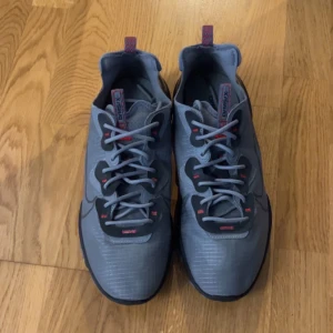 Nike React grå sneakers med röda detaljer - Nike React sneakers i grått med svarta och röda detaljer. Ovandel i syntetmaterial med diskret randigt mönster.