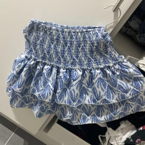 Blåvit volangkjol med mönster - Supersöt kort kjol i blått och vitt med volanger och elastisk midja. Kjolen har ett snyggt bladmönster och är perfekt för varma dagar. Lätt och luftig känsla, passar till sneakers eller sandaler. Denna kjol är i storlek XS men passar för Storlek M