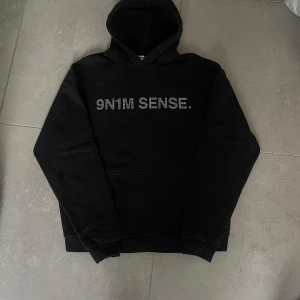 Svart hoodie 9N1M SENSE med strass - Svart hoodie från 9N1M SENSE med glittrig strass-text framtill. Klassisk modell med huva och magficka, ribbade muddar och relaxed fit. Perfekt för dig som gillar streetstyle och vill sticka ut med snygga detaljer.