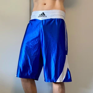 Blå Adidas träningsshorts med vita detaljer - Snygga blå boxningsshorts från Adidas med bred vit resårmidja och klassiska vita ränder längs sidorna. Långa ben och loose fit, perfekt för sport eller gym. Materialet är glansigt och lätt, troligen polyester. Adidas-logga framtill på midjan. Storlek XL men passar även M och L utmärkt!