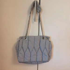 Quiltad randig axelväska med kedja ZARA - Säljer en quiltad axelväska i ljusblått och vitt med tunna ränder. Väskan har silverfärgade kedjehandtag och magnetlås. Mjuk och vadderad känsla, perfekt för att bära allt du behöver. Snygg och unik design som sticker ut.