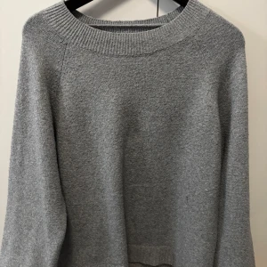 Grå stickad tröja - Mysig grå stickad tröja med rund halsringning och ribbade muddar. Perfekt för lager-på-lager och enkel att matcha med både jeans och kjol. Tröjan har en klassisk passform och långa ärmar, vilket gör den superbekväm under kyliga dagar.