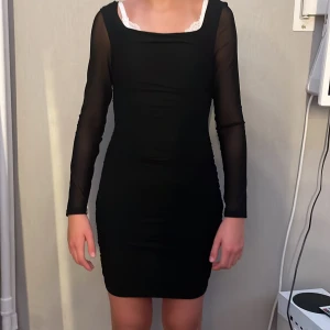 Svart tight meshklänning - Snygg svart kortklänning med tight passform och långa meshärmar. Klänningen har en fyrkantig ringning och är tillverkad i stretchigt syntetmaterial. Perfekt för dig som gillar en stilren look med lite edge.