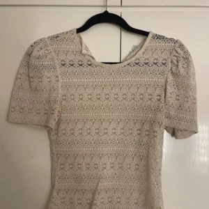 Vit spetsblus från Vila XS - Supersöt vit spetsblus från Vila i storlek XS. Blusen har korta puffärmar och ett detaljerat spetsmönster över hela plagget. Perfekt för dig som gillar romantisk och feminin stil. Rund halsringning och figurnära passform.