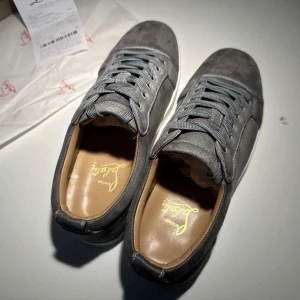 Grå mockasneakers från Louboutin - Snygga grå sneakers från Christian Louboutin i mjuk mocka med klassisk röd sula och vita kanter. Skorna har rund tå, platt sula och grå snörning. Insidan är i beige läder för extra komfort. Perfekta för dig som vill sticka ut med exklusiv stil.