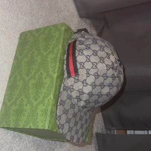 Gucci keps med GG-mönster S - Exklusiv Gucci keps i beige och mörkblått GG-monogram, tillverkad i bomullscanvas. Kepsen har klassisk böjd skärm och ikoniska röda och gröna ränder på sidan. Perfekt accessoar för dig som vill sticka ut med lyxig streetstyle. 