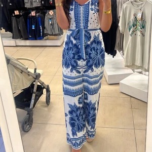 Vit och blå byxdress med blommor - Snygg byxdress i vitt med stora blå blommor och mönster. Vida ben och knytband i midjan ger en avslappnad och trendig look. Perfekt för sommardagar och festivaler. Lätt och luftig, med breda axelband och v-ringning framtill.