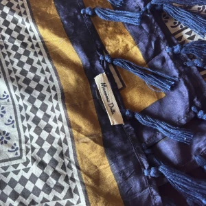 Mönstrad halsduk från Massimo Dutti - Snygg halsduk från Massimo Dutti med blå och senapsgula kanter, vita och svarta geometriska mönster samt blå tofsar längs kanten. Perfekt accessoar för att lyfta din outfit med lite extra färg och stil.