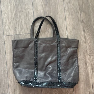 Grå skinnväska med paljettdetaljer - Stor grå handväska från Vanessa Bruno i äkta skinn med svarta paljetter längs handtag och botten. Väskan har innerficka med dragkedja och rymligt fack. Perfekt för dig som gillar en stilren look med lite extra bling.