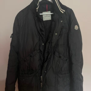Svart dunjacka från Moncler - Snygg svart vintage dunjacka från Moncler med klassisk logga på ärmen och coola detaljer som dragkedja och knappar framtill. Jackan har hög krage med randig ribbad kant och flera fickor. Perfekt för dig som vill ha en stilren och varm jacka.