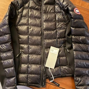 Svart dunjacka från Canada Goose - Snygg svart dunjacka från Canada Goose med quiltad design och klassisk logga på ärmen. Jackan har dragkedja framtill, två sidofickor med dragkedja och är tillverkad i glansigt material. Perfekt för kalla dagar och riktigt stilren look.