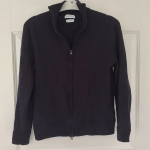 Svart merinoullskofta från These Glory Days - Stilren svart kofta i merinoull från These Glory Days. Modellen har hel dragkedja framtill, hög krage och långa ärmar. Perfekt för lager-på-lager och ger en clean look till din outfit. Inuti står set storlek L men den passar någon som har S/XS.