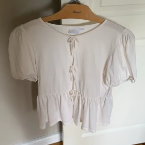 Vit blus med knyt och puffärm - Supersöt vit blus från Gina Tricot med puffiga korta ärmar och flera knyt framtill. Blusen har en volang nertill som ger en romantisk vibe och är gjord i mjuk bomull. Perfekt för dig som gillar en feminin och trendig stil.