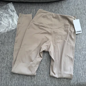 Beige leggings från Stronger - Supersnygga beige leggings från Stronger i storlek XS. De är högmidjade och har en tight passform som sitter perfekt vid träning. Tillverkade i ett mjukt och stretchigt syntetmaterial som känns skönt mot huden. Diskret logga framtill i samma färg.
