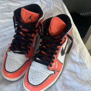 Nike Air Jordan 1 High Orange/White - Säljer ett par Nike Air Jordan 1 High i orange, vitt och svart. Skorna har klassisk hög siluett, svart swoosh, svarta skosnören och perforerad tå. Tillverkade i skinn med kontrasterande färgblock och ikonisk Jumpman-logga på plösen.