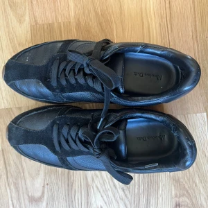 Svarta sneakers från Massimo Dutti - Stilrena svarta sneakers från Massimo Dutti med ovandel i skinn och mocka, snygga svarta snören och diskret logga i guld på hälen. Skorna har en platt sula med bruna och vita detaljer och rund tå. Perfekta för en clean och modern look.