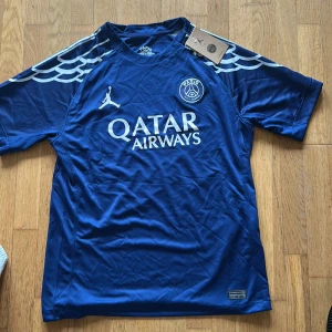 Blå PSG fotbollströja från Jordan - Snygg blå Paris Saint-Germain fotbollströja från Jordan med Qatar Airways-tryck på bröstet. Tröjan har korta ärmar och coola vita vingmönster på axlarna. Tillverkad i lätt och ventilerande material, perfekt för match eller träning. 