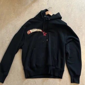 Svart hoodie med blommigt smiley-motiv - Svart hoodie med stor blommig smiley-applikation på ryggen och unik broderad detalj framtill. Oversized. 