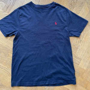 Mörkblå t-shirt från Polo Ralph Lauren - Mörkblå t-shirt från Polo Ralph Lauren med röd  logga. Storlek 10-12 år, 150 