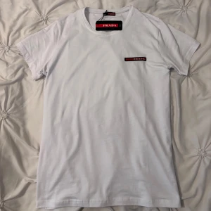 Prada T-shirt - Vit t-shirt från Prada i mjuk bomull med klassisk passform. T-shirten har korta ärmar och en stilren röd och svart Prada-logga på bröstet. Perfekt basic med exklusiv känsla och snygg rund halsringning.
