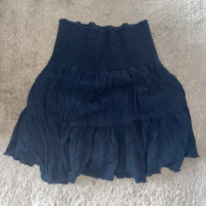 Mörkblå volangkjol från 157 - Supersöt mörkblå kortkjol från 157 med bred smock i midjan och volanger nertill. Kjolen är tillverkad i 100% viskos vilket gör den lätt och skön att bära. Perfekt för dig som gillar en feminin och avslappnad stil.