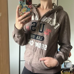 Brun hoodie med tryck  - Säljer en brunmelerad hoodie från Tindra med stora broderade bokstäver och tryck på framsidan. Tröjan har huva med snörning, dragkedja och ribbade muddar. Perfekt för en chill och sportig look. Använd fåtal gånger! Storlek S- 36/38🤎