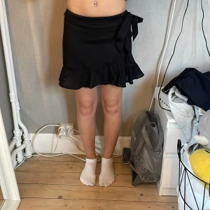 Svart omlottkjol med volang från SHEIN - Supersöt svart kort omlottkjol från SHEIN i lätt och flowigt material. Kjolen har en bred volang längs nederkanten och knyts i sidan med en stor rosett. Perfekt för dig som gillar en feminin och trendig look.