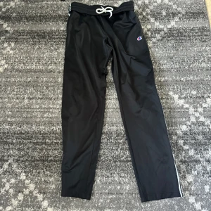 Champion byxor - Svarta träningsbyxor från Champion med vita och svarta ränder längs benen och logga på sidan. Byxorna har snörning i midjan och är gjorda i polyester. Perfekta för chill eller sportiga dagar.