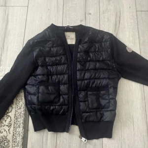 Moncler cardigan  - Snygg moncler cardigan pufferjacka från Moncler med quiltad framsida och två fickor. Jackan har långärmade stickade ärmar och ribbade muddar. och har klassisk Moncler-logga på ärmen. Perfekt för dig som vill ha en stilren och exklusiv look. Obs : dragkedjan är borta!