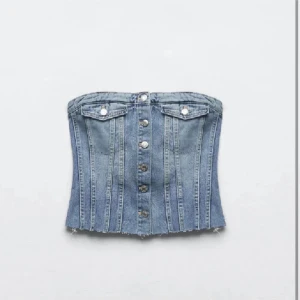 Jeans korsett-topp med knappar - Säljer en snygg jeans korsett-topp med knappar framtill och två dekorativa fickor vid bysten. Toppen har en croppad passform och råa kanter nertill för en cool look. Perfekt att styla med höga byxor eller kjol.