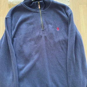  Polo Ralph Lauren Half Zip - Säljer en marinblå långärmad tröja från Polo Ralph Lauren med halv dragkedja och klassisk röd logga broderad på bröstet. Tröjan är i bomull och har en hög krage som ger en clean och stilren look. Perfekt för lager-på-lager under kyliga dagar.