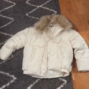 Beige pufferjacka med fuskpäls Missguided - Säljer en beige pufferjacka från Missguided med stor, fluffig fuskpäls på kragen. Jackan har dragkedja och tryckknappar framtill, samt två stora fickor. Perfekt för kalla dagar när du vill vara både varm och snygg.