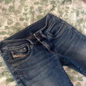 Blå jeans från Diesel - Snygga blå jeans från Diesel med klassisk femficksdesign. Jeansen har slitningar och ljusare partier framtill, samt detaljer i rött vid knappar och sömmar. Tillverkade i jeansmaterial med en cool tvättad look. Innerbenslängden är 77cm, de är lite slitna nertill. Tveka inte med att fråga om bilder 💕