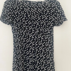 Blommig kortklänning från Lindex - Supersöt svart kortklänning från Lindex med vita små blommor över hela tyget. Klänningen har v-ringning, korta ärmar och en lätt figursydd passform. Perfekt för dig som gillar en feminin och trendig stil.