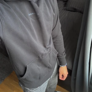 Svart hoodie från Calvin Klein Jeans - Svart hoodie från Calvin Klein Jeans med diskret logga på bröstet och snyggt tryck på huvkanten. Klassisk känguruficka framtill och ribbade muddar. Perfekt för en chill och stilren look.