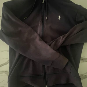 Svart hoodie från Polo Ralph Lauren - Svart hoodie från Polo Ralph Lauren med dragkedja och klassisk broderad logga på bröstet. Tröjan har huva med snörning och långa ärmar. Perfekt för en chill och stilren look. Materialet är mjuk bomull som känns skönt mot huden.