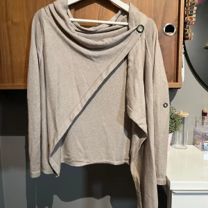 Beige omlottkofta med knappdetalj - Snygg beige omlottkofta med lång ärm och cool knappdetalj vid axeln. Koftan har en avslappnad passform och är tillverkad i ett mjukt, stickat material. Perfekt för lager-på-lager och chill dagar. 