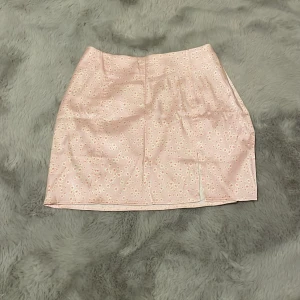 Rosa blommig kjol från Zara - Supersöt rosa kortkjol från Zara med små vita och orange blommor. Kjolen har en rak passform och en liten slits framtill som ger en extra touch. Perfekt för dig som gillar en feminin och trendig stil.