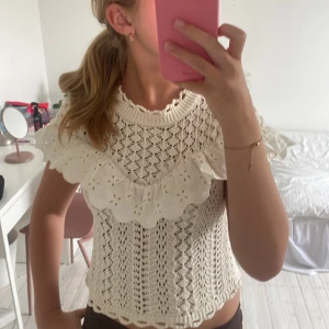Vit stickad blus med volang från Zara - Superfin vit stickad blus från Zara med broderad volangdetalj över axlarna. Toppen har korta ärmar, högt ribbat halslinne och fint hålmönster i bomull. Jätte snygg till ett par jeans. Fint skick, utan defekter.
