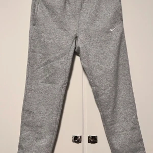 Nike grå mjukisbyxor   - Snygga grå mjukisbyxor från Nike med klassisk vit swoosh-logga på låret. Byxorna har elastisk midja, raka ben och är tillverkade i mjukt bomullsmaterial som håller dig varm. Perfekt för att ha ute på hösten eller på vintern , med fickor på sidorna och en bakficka.Längd 95cm och opening 18cm 