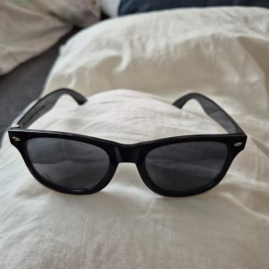 Svarta solglasögon från BE JOYND - Snygga svarta solglasögon från BE JOYND med klassisk wayfarer-form. Bågarna är blanka och tillverkade i plast, med mörka glas och silvriga detaljer vid fronten. Perfekta för att ge en cool vibe till din stil.