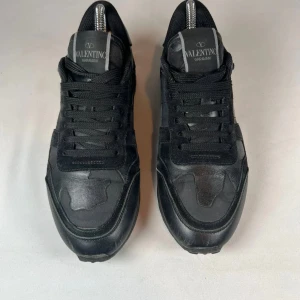 Valentino Garavani Rockrunner sneakers - Svarta Valentino Garavani Rockrunner sneakers med coola nitar på hälen. Skorna har en mix av skinn och mocka, samt textilpaneler för en lyxig känsla. Klassisk snörning och diskret camo-mönster ger en edgy vibe. Perfekta för dig som gillar stilrena men unika sneakers.