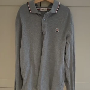 Grå långärmad pikétröja Moncler - Snygg grå långärmad pikétröja från Moncler med klassisk krage och knäppning framtill. Tröjan har diskret Moncler-logga på bröstet och röd-vit detalj på kragen. Perfekt för dig som gillar stilren och sportig look.