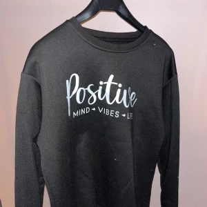 Svart sweatshirt med tryck 'Positive' - Svart sweatshirt med vitt tryck på bröstet där det står 'Positive Mind Vibes Life'. Tröjan har rund halsringning och långa ärmar. Perfekt för dig som gillar en enkel men peppig stil. Materialet känns mjukt och bekvämt, troligen bomull eller polyester.