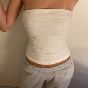 Vit bandeau tubtopp med struktur - Säljer en vit bandeau tubtopp med snyggt strukturerat tyg. Toppen är axelbandslös och har en tight passform som sitter snyggt på kroppen. Perfekt för varma dagar eller när du vill ha en clean och stilren look.