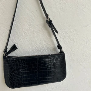 Svart axelväska i croco-imitation - Snygg svart axelväska med croco-mönstrad yta och justerbart axelband med silverfärgat spänne. Finns även ett fack med dragkedja invändigt i väskan! 
