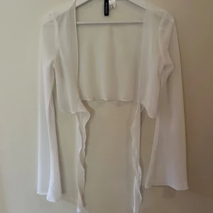 Vit transparent kofta från H&M Divided - Säljer en supertunn och transparent vit kofta från H&M Divided. Den är croppad, har långa ärmar och öppet framtill med vågiga kanter. Perfekt att slänga över ett linne eller klänning för en lätt och luftig look.