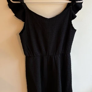 Svart kortklänning från H&M - Supersöt svart kortklänning från H&M med smockat tyg och volangdetaljer vid axlarna. Klänningen är ärmlös och har en lätt v-ringad halsringning. Perfekt för dig som gillar en enkel men trendig look.